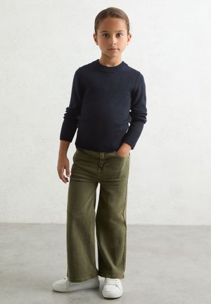 Barn står iført en navyblå sweater, olivengrønne vidde bukser og hvide sneakers med den ene hånd i lommen mod en ensfarvet baggrund.