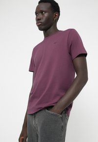 Tricou cu mânecă scurtă, guler rotund, de culoare mov închis, din bumbac, având un mic detaliu cu logo pe piept. Asortat cu blugi gri.