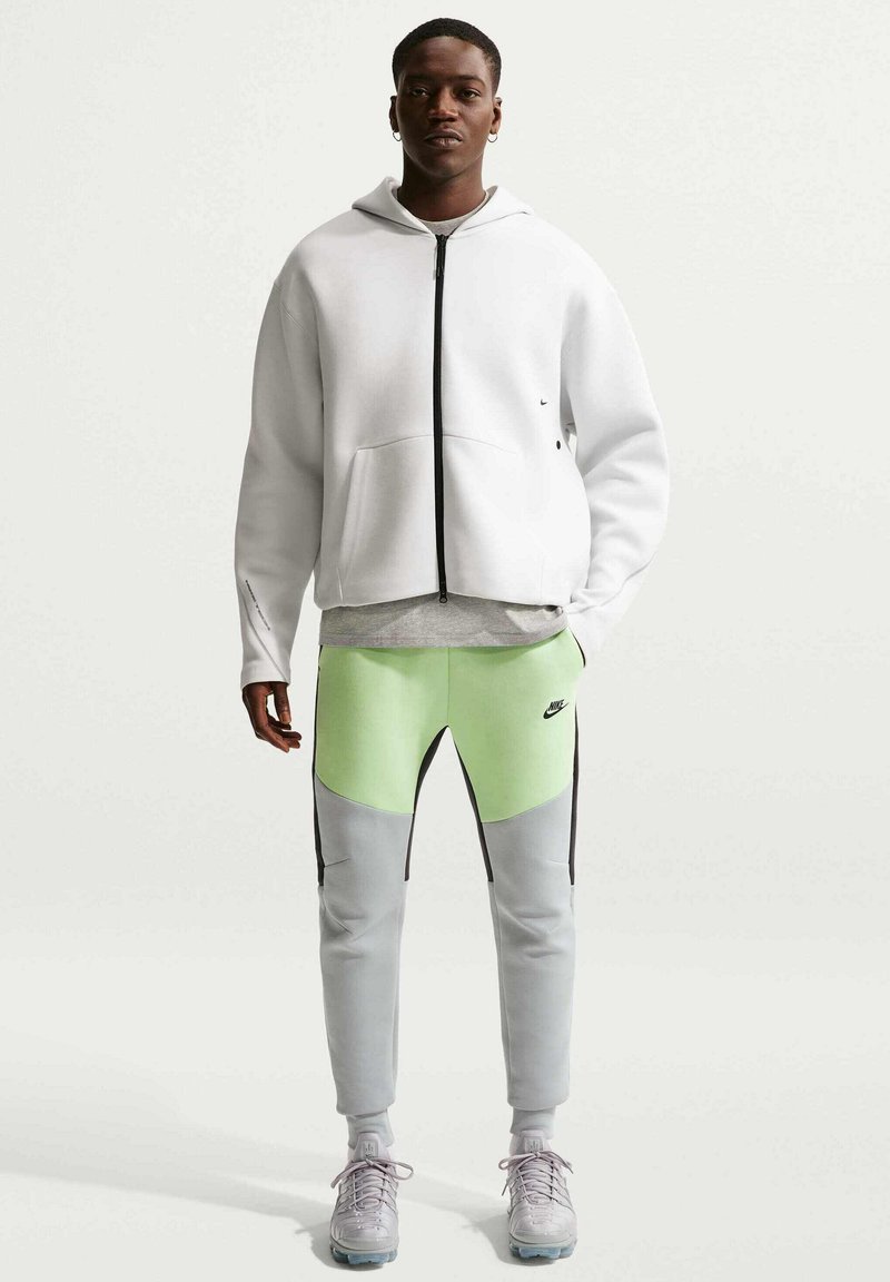 Mand iført hvid hættetrøje med lynlås, grå og neongrønne Nike-bukser og grå sneakers, stående foran en ensartet lys baggrund.