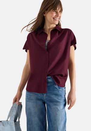 Smilende kvinde med skulderlangt brunt hår iført en kortærmet, mørkerød knap-op skjorte og blå jeans, der holder en denim tote-taske.