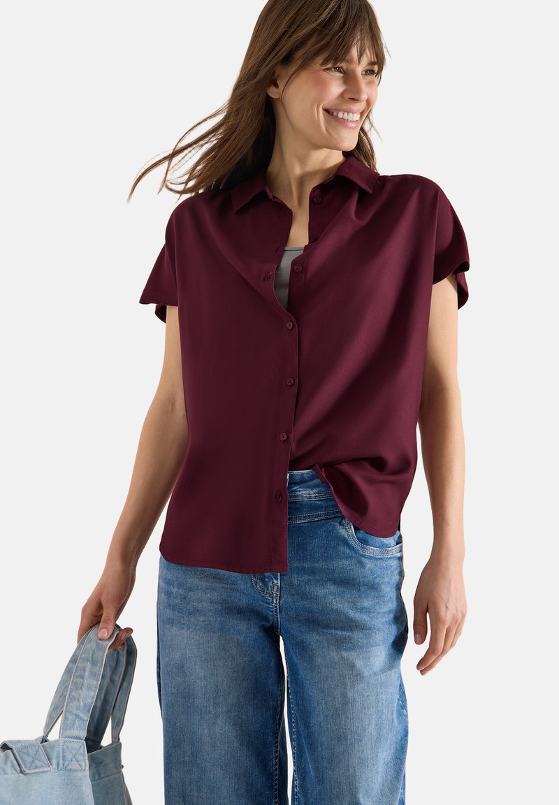 Femme souriante aux cheveux bruns mi-longs portant une chemise bordeaux à manches courtes et un jean bleu, tenant un sac fourre-tout en denim.