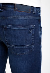Vaqueros de mezclilla azul oscuro con un parche de cuero texturizado de la marca, que cuentan con detalles en los bolsillos traseros y costuras en contraste en la cinturilla.