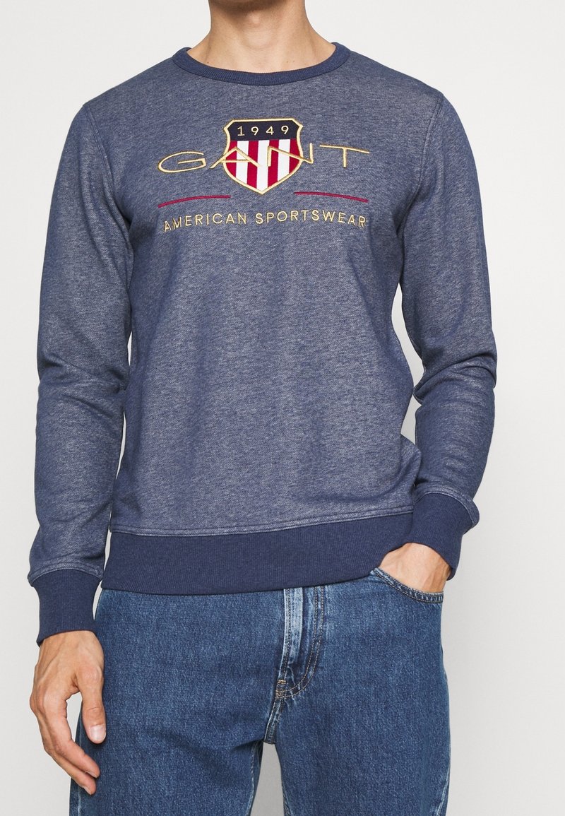 Homme portant un sweat-shirt bleu marine avec le logo "GANT 1949 American Sportswear" et un jean bleu, main dans la poche, debout devant un fond blanc.