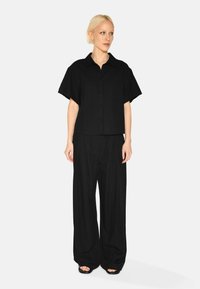 Black, short-sleeve button-up shirt with a pocket, paired with loose-fitting black trousers. Smooth texture, minimalistic design, no patterns.
