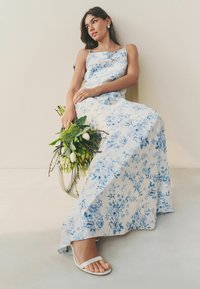 Rochie lungă, fluidă, din material alb cu imprimeu floral albastru. Modelul ține un buchet de verdeață. Sandale albe minimaliste. Design simplu.