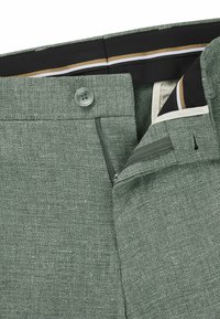 Zielone spodnie tkane o teksturowanym wykończeniu, z zapięciem na guzik, kieszenią na zamek oraz kontrastującym wewnętrznym pasem ze smugami.