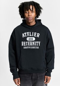 Must mustriga kapuutsiga särk, millel on valge tekst: "ATELIER RETERNITY 2018 LOOVJUHT." Omab eesmist taskut ja soonikkoes varrukaid.