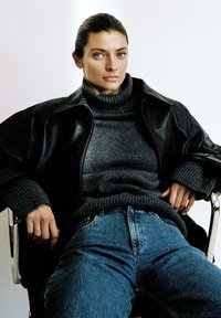 Schwarze Lederjacke, grauer Rollkragenpullover und blaue Jeans. Modell sitzt auf einem Stuhl, mit schulterlangen dunklen Haaren und neutralem Ausdruck.