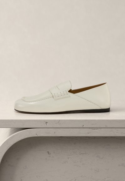 Λευκό δερμάτινο loafers χωρίς κορδόνια με λεπτομέρεια ραφιών penny σε υφή ανοιχτό γκρίζο ράφι πάνω σε απλό μπεζ φόντο.