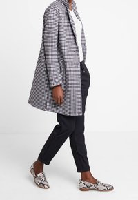 Zwart-wit geruite oversized jas, witte blouse eronder, zwarte tapered broek en loafers met slangenprint, waarmee verschillende texturen en patronen worden getoond.