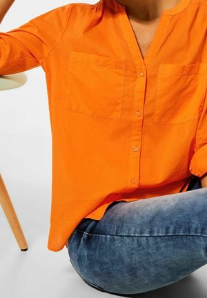 Donna che indossa una camicia arancione brillante con tasche sul petto e jeans blu, seduta accanto a una gamba di sedia in legno chiaro su pavimento bianco.