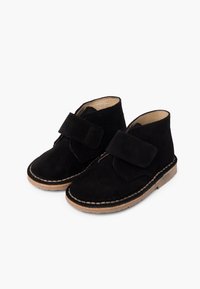 Bottines en daim noir pour enfants, avec un bout arrondi, une bande Velcro et une semelle contrastante de couleur marron clair avec des détails de couture visibles.