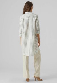 Camicia a maniche lunghe a righe bianca e grigia chiara, con colletto a camicia e maniche arrotolate, abbinata a pantaloni a gamba larga color crema.
