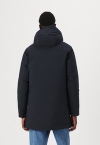 Parka imbottita di un blu scuro navy, con un grande cappuccio, taglio dritto e texture liscia. Presenta un retro leggermente più lungo e dettagli minimi.