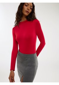Donna in una maglietta rossa aderente a maniche lunghe e una gonna grigia alta in vita con uno spacco frontale, in posa con un braccio dietro la schiena.