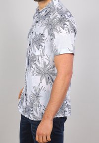 Mies, joka käyttää lyhythihaista nappipaitaa, jossa on musta-harmaat trooppiset lehtikuviot, yhdistettynä tummaansinisiin farkkuihin, nähtynä sivulta.