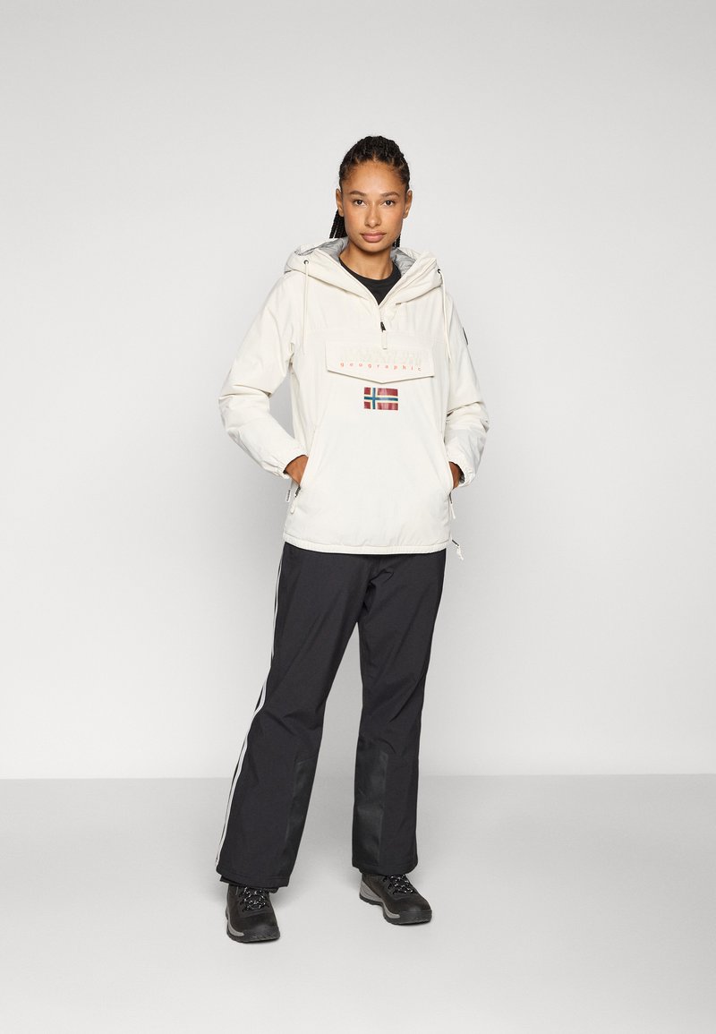 Anorak blanco con un bolsillo frontal, que presenta un diseño de la bandera de Noruega en rojo y verde. Combinado con pantalones de outdoor negros y botines negros resistentes.