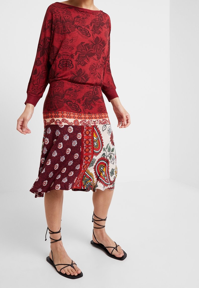 Femme portant un haut rouge à motifs floraux et une jupe patchwork multicolore avec des imprimés cachemire et floraux, associée à des sandales plates noires à lacets.