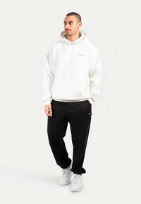 Felpa bianca con cappuccio e tasca a marsupio con logo ricamato, abbinata a pantaloni neri e sneakers bianche, modello in piedi su sfondo neutro.