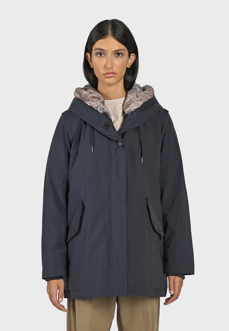 Parka bleu marine avec une capuche doublée en fausse fourrure, deux poches avant et des ajustements à toron. Le matériau semble lisse et durable.