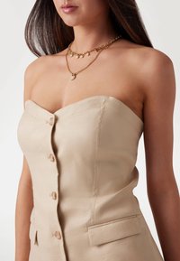 Abito beige senza spalline con un corpetto aderente, bottoni frontali e tasche laterali. Collane dorate a strati con ciondoli accentuano il collo.