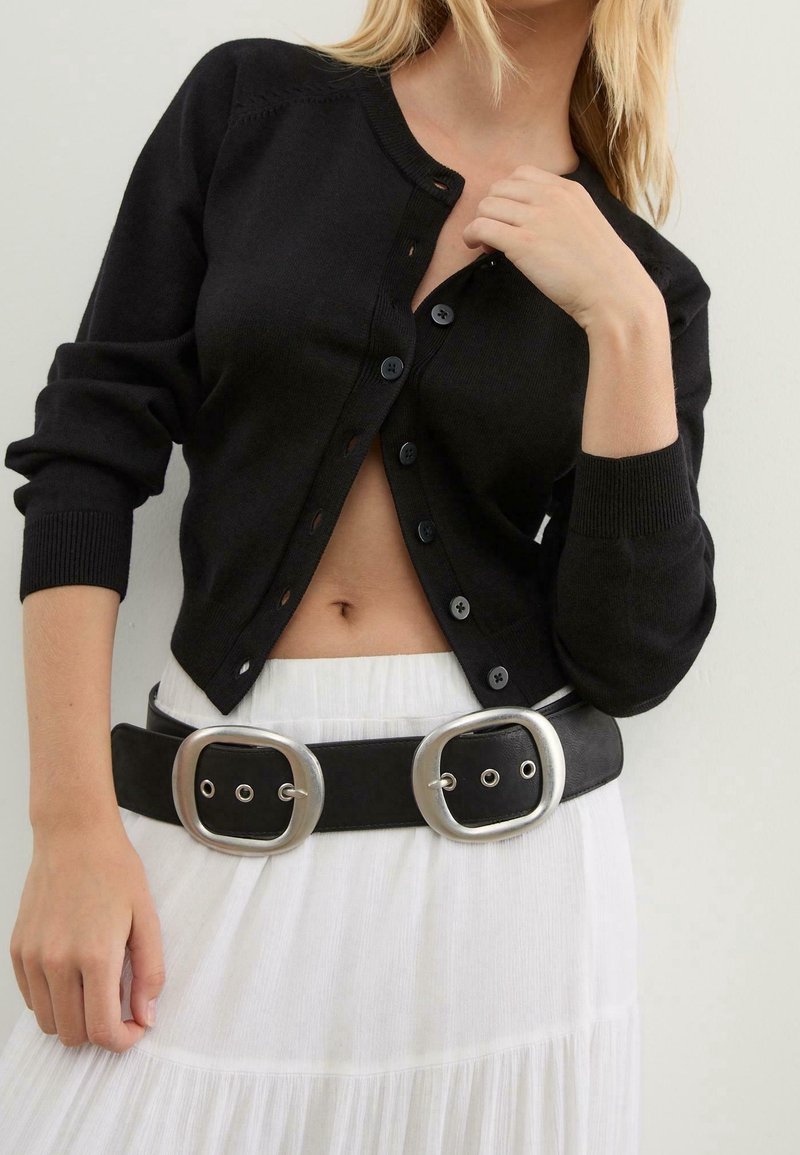 Femme portant un cardigan noir boutonné, une jupe blanche et une ceinture noire avec deux grandes boucles argentées, main près de la clavicule.