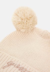 Cappello invernale lavorato a maglia di colore crema con un grande pompon in cima e nome del marchio ricamato sul risvolto.