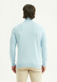 Pull à col roulé bleu clair en tissu doux, design ajusté, manches longues, poignets et ourlet côtelés. Porté avec un pantalon de couleur claire.