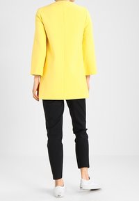 Abrigo amarillo de corte recto, con mangas hasta el codo y una costura en la parte de atrás, combinado con pantalones negros y zapatillas blancas.