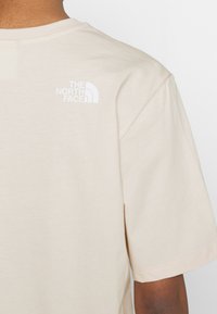 T-shirt in cotone beige con colletto rotondo e maniche corte. Sul lato sinistro della schiena è presente un logo bianco de "The North Face". Texture morbida.