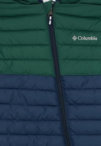 Zaļš un tumši zils quilted jaka ar centrālo rāvējslēdzēju un Columbia logotipu labajā augšējā krūtī.