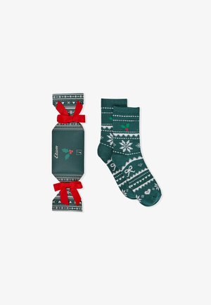 Calzini invernali verdi con disegni bianchi di fiocchi di neve e agrifoglio, abbinati a un cracker regalo con nastri rossi e un motivo festivo.