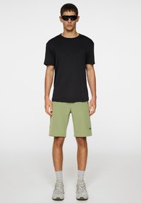 Svart kortärmad t-shirt och ljusgröna shorts, kombinerat med grå sneakers och mörka solglasögon; avslappnad sportklädsel med en ledig passform.