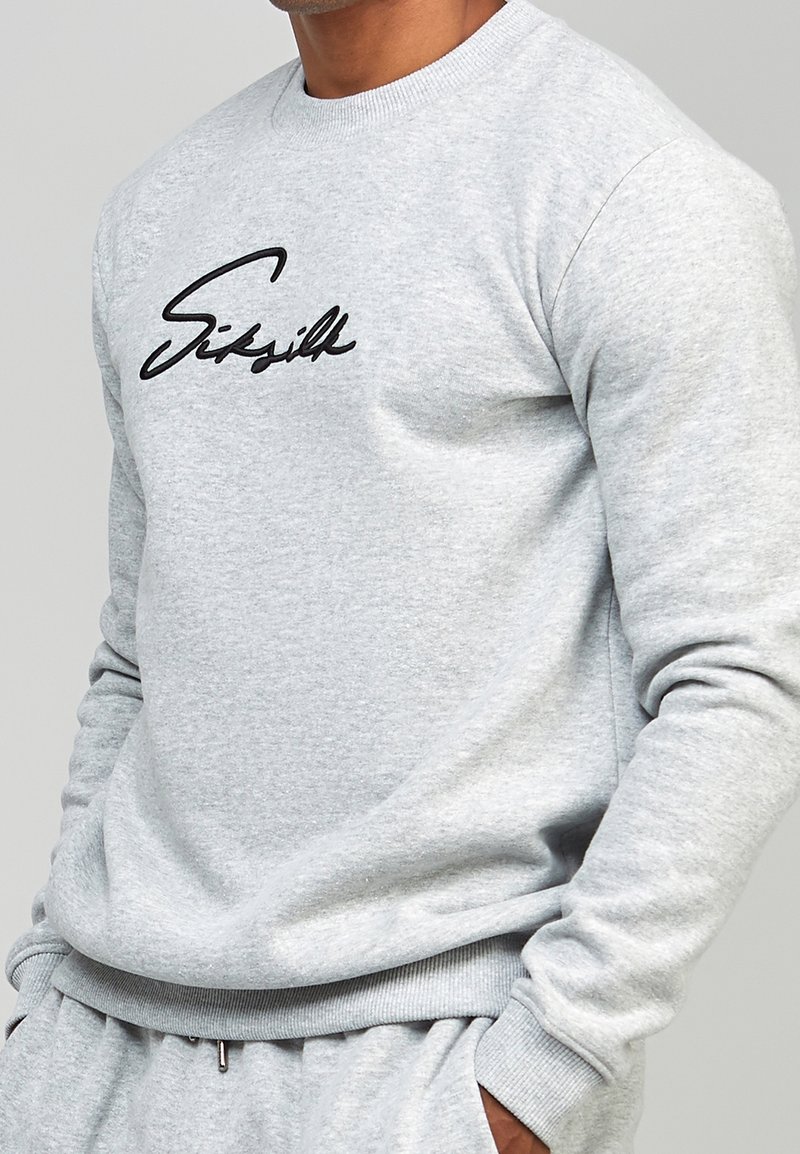 Homme portant un sweat-shirt gris clair et un pantalon de jogging assorti, avec un logo "SikSilk" noir en écriture cursive sur le devant du sweat-shirt.