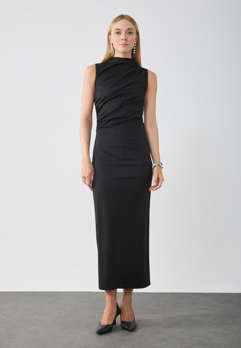 Black, fitted, sleeveless dress with gathered fabric on the bodice, reaching below the knee. Worn with black high-heeled shoes.