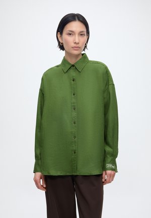 Mujer con pelo corto y negro que lleva una camisa verde holgada abotonada y pantalones marrones oscuros sobre un fondo blanco liso.