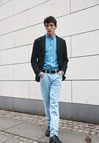 Joven caminando con las manos en los bolsillos, vestido con una blazer negra, camisa azul abotonada, jeans claros y zapatos negros en una acera.