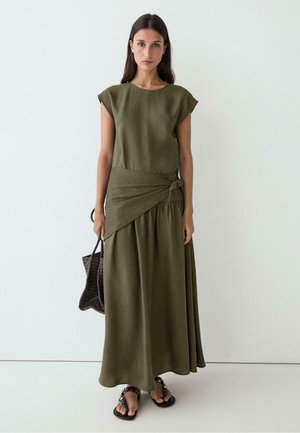 Donna che indossa un vestito lungo senza maniche verde oliva con dettaglio annodato in vita, tiene una borsa intrecciata e indossa sandali neri, in piedi contro un muro bianco.