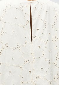 Λευκό ύφασμα με μεγάλη κεντητή λουλουδένια διάτρητη υφή και μικρό στρογγυλό κουμπί πάνω από κάθετη σχισμή σε σχήμα κλειδιού.