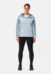 Lichtblauwe zip-up jas met een capuchon en contrasterende donkere mouwen, gecombineerd met zwarte leggings en zwarte wandelschoenen.