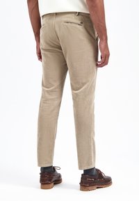 Beige Cordhosen mit geradem Schnitt, dezenten vertikalen Streifen und Detail an der Gesäßtasche, kombiniert mit dunkelbraunen Schuhen und grauen Socken.