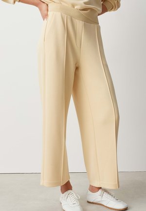 Persoon die beige wijde broek draagt met een elastische tailleband en witte sneakers, staand op een lichtgrijze vloer.
