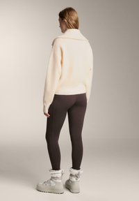 Nő krémszínű, bő pulóverben, sötétbarna leggingsben és fehér, bélelt téli csizmában, a kamerának háttal áll.