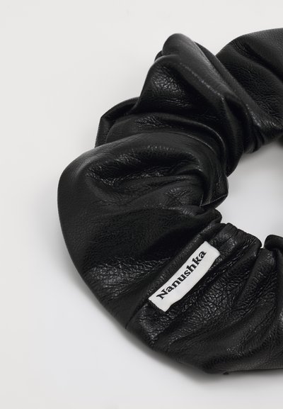 Melnā ādas scrunchie ar teksturētu virsmu un baltu etiķeti ar "Nanushka" zīmolu. Apaļa forma ar savelkamiem elementiem.