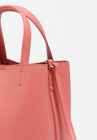 Bolso de cuero rosa con textura suave, asas cortas y una correa de hombro desmontable. Presenta costuras visibles y un detalle de botón.