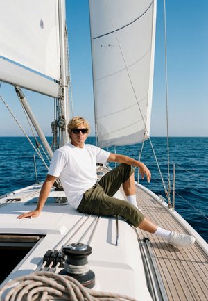 Un jeune homme en lunettes de soleil se détend sur le pont d'un voilier aux voiles blanches sur une mer bleue calme sous un ciel dégagé.