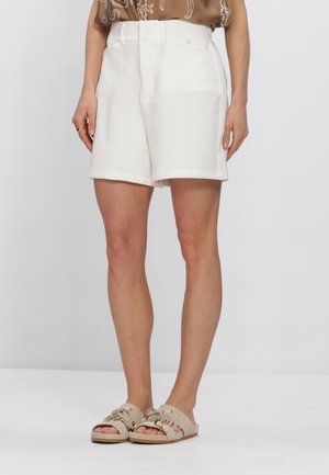 Vrouw draagt witte op maat gemaakte korte broek en beige sandalen met franjes aan de bandjes tegen een effen grijze achtergrond.