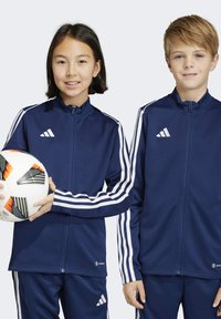 Marineblaue Sportjacken mit weißen drei Streifen, getragen von zwei Kindern. Eines hält einen gemusterten Fußball.