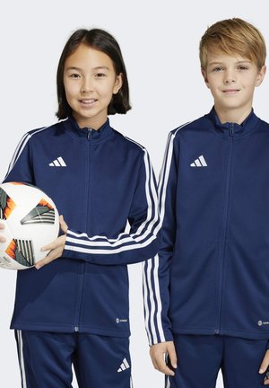 Veste de sport zippée bleu marine avec des accents blancs à trois rayures, portée par deux enfants. L'un tient un ballon de football à motifs.