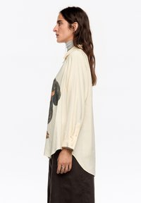 Camisa oversize color crema con un estampado gráfico en el frente, mangas largas, puños con botones y dobladillo curvado, combinada con un cuello alto gris.